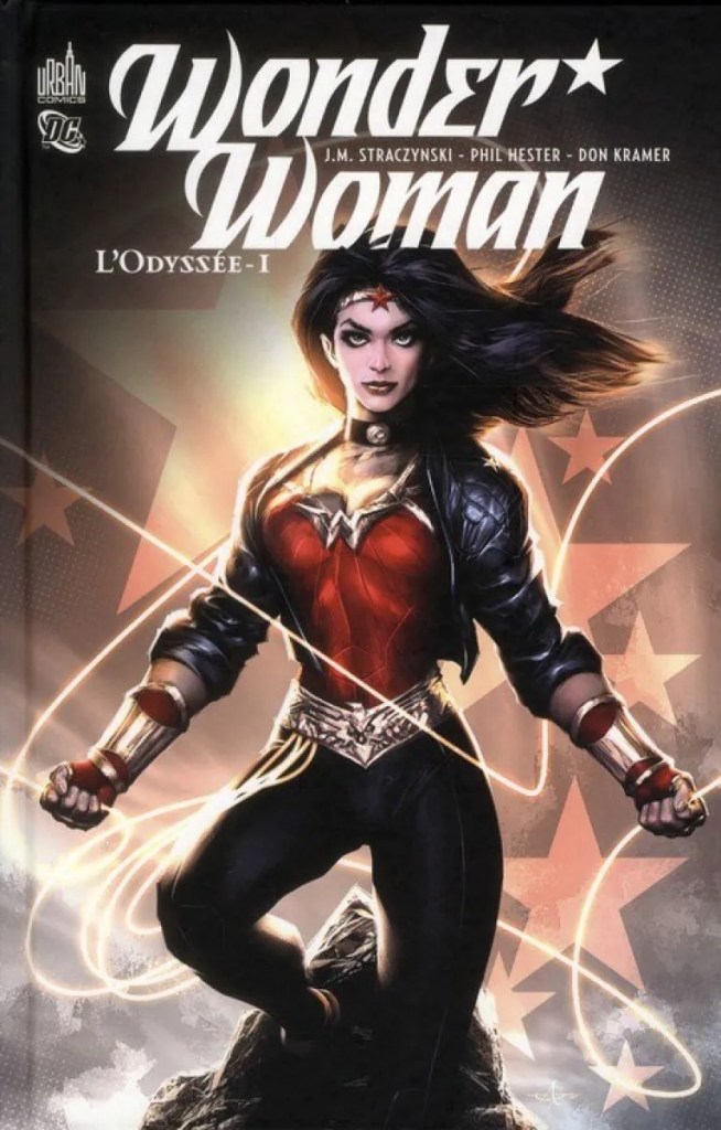 Couverture du comics Wonder Woman - L'Odysée - 1
On y voit Wonder Woman debout le torse "bombée", puissante, vêtu de son mythique bustier rouge, une veste en cuir noire, et un pantalon noir. Derrière on devine les étoiles du drapeau américain.